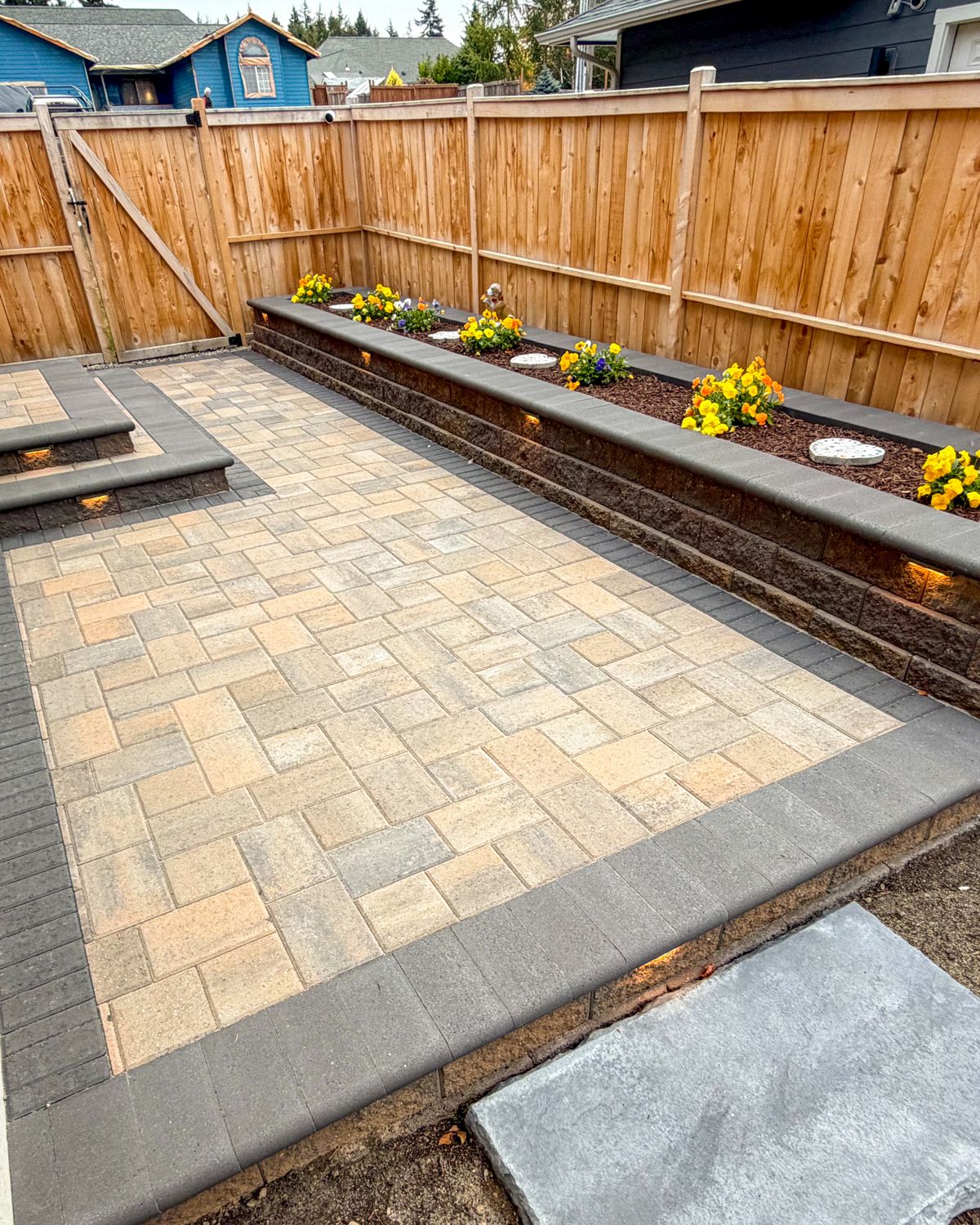 Paver patio wrapped by a lit raised block planter with yellow flowers